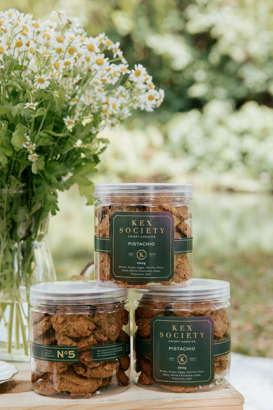 Three clear jars of 250g Kex Society roasted pistachio cookies with cookies displayed in front of the jars. The setting appears to be outdoors with natural lighting and some greenery in the background.