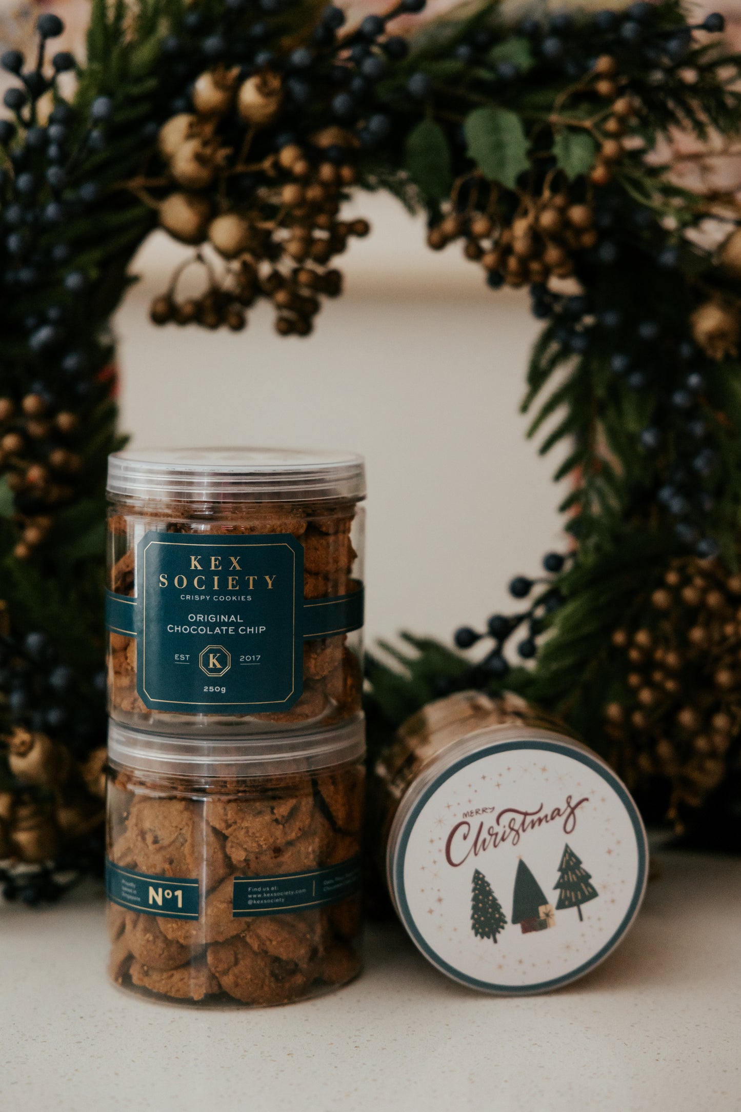 Two stacked containers of Kex Society chocolate chip cookies with a Christmas-themed lid in front of a decorative wreath.