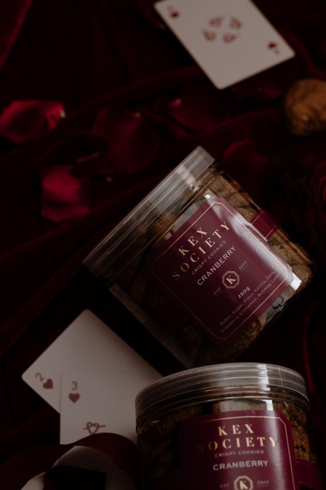 2 Kex Society 250g bottles of crispy cranberry cookies displayed among playing cards on a velvet red cloth with rose petals around.