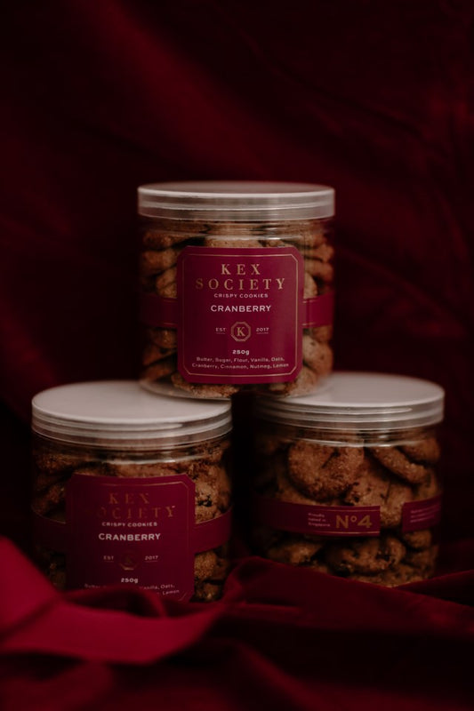 Three jars of 'KEX SOCIETY CRANBERRY' cookies placed against a dark, red backdrop. The jars are clear, allowing the cookies inside to be visible, and are sealed with customized lids.