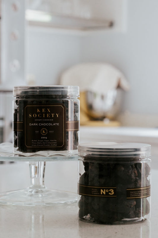 Two clear  Kex Society 250g jars on a kitchen counter filled with triple dark chocolate cookies, each jar with a different label indicating the product is 'No. 3 Dark Chocolate Cookies'.