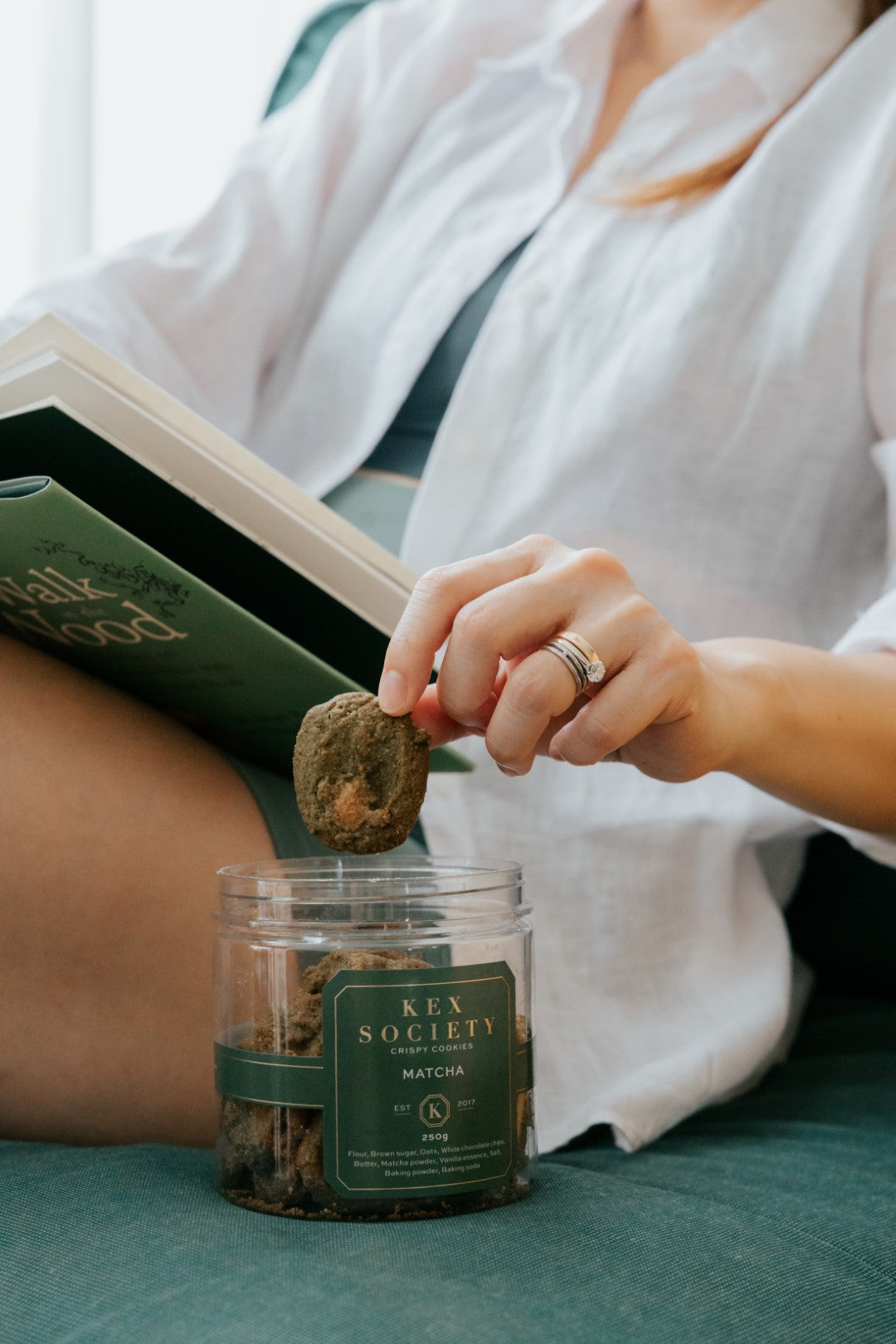 A lady in casual linen clothing and active wear with a book on her lap. She's reaching out for a Kex Society Matcha White Chocolate cookie from an opened half filled jar.