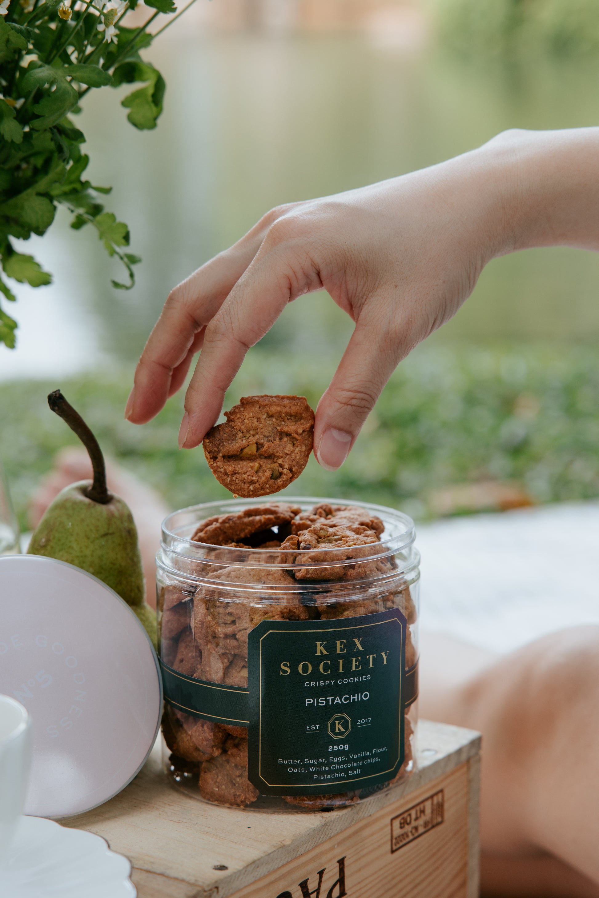 An open bottle of 250g Kex Society Roasted Pistachio cookies with a hand picking out a single cookie. Bottle is placed on a wooden carton with flowers and a pear next to it. Background is outdoor greenery.