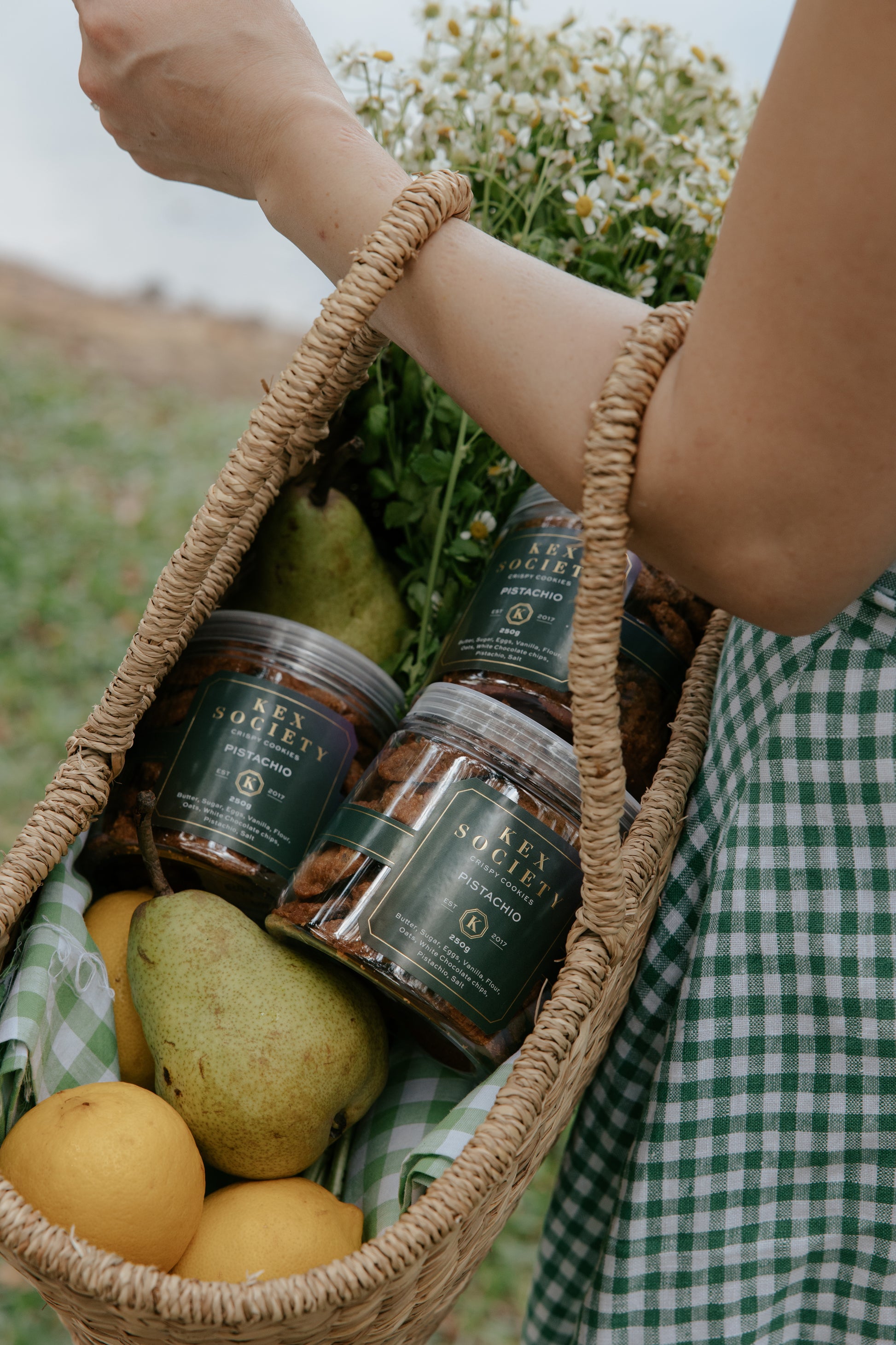 A woven basket held by an arm, inside are 3 bottles of 250g crispy Kex Society Roasted Pistachio cookies with lemons, pears and a bouquet of flowers.