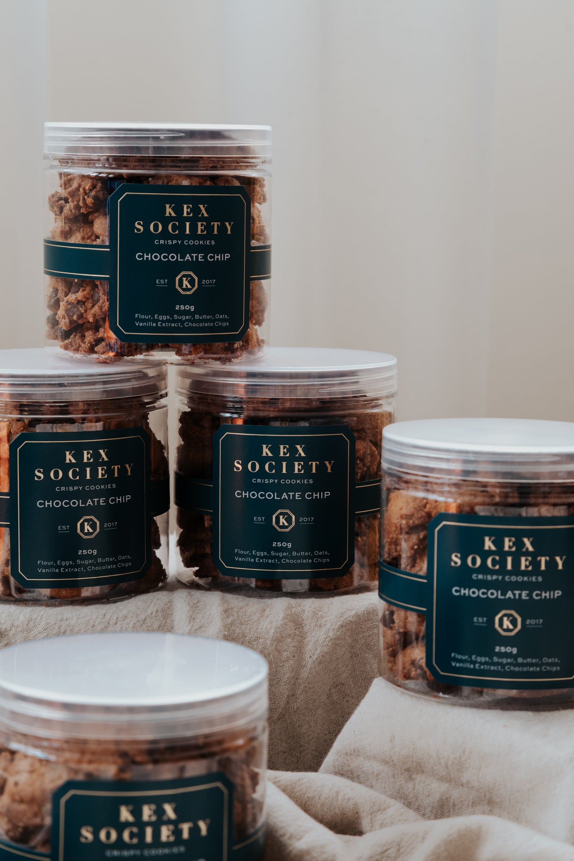 250g Kex Society bottles containing crispy chocolate chip cookies. Stacked in an aesthetic way with a neutral background