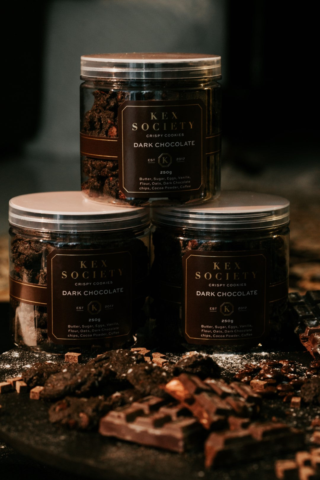 3 bottles of  Kex Society's crispy Triple dark chocolate cookies displayed among scattered cookies and blocks of dark chocolate.