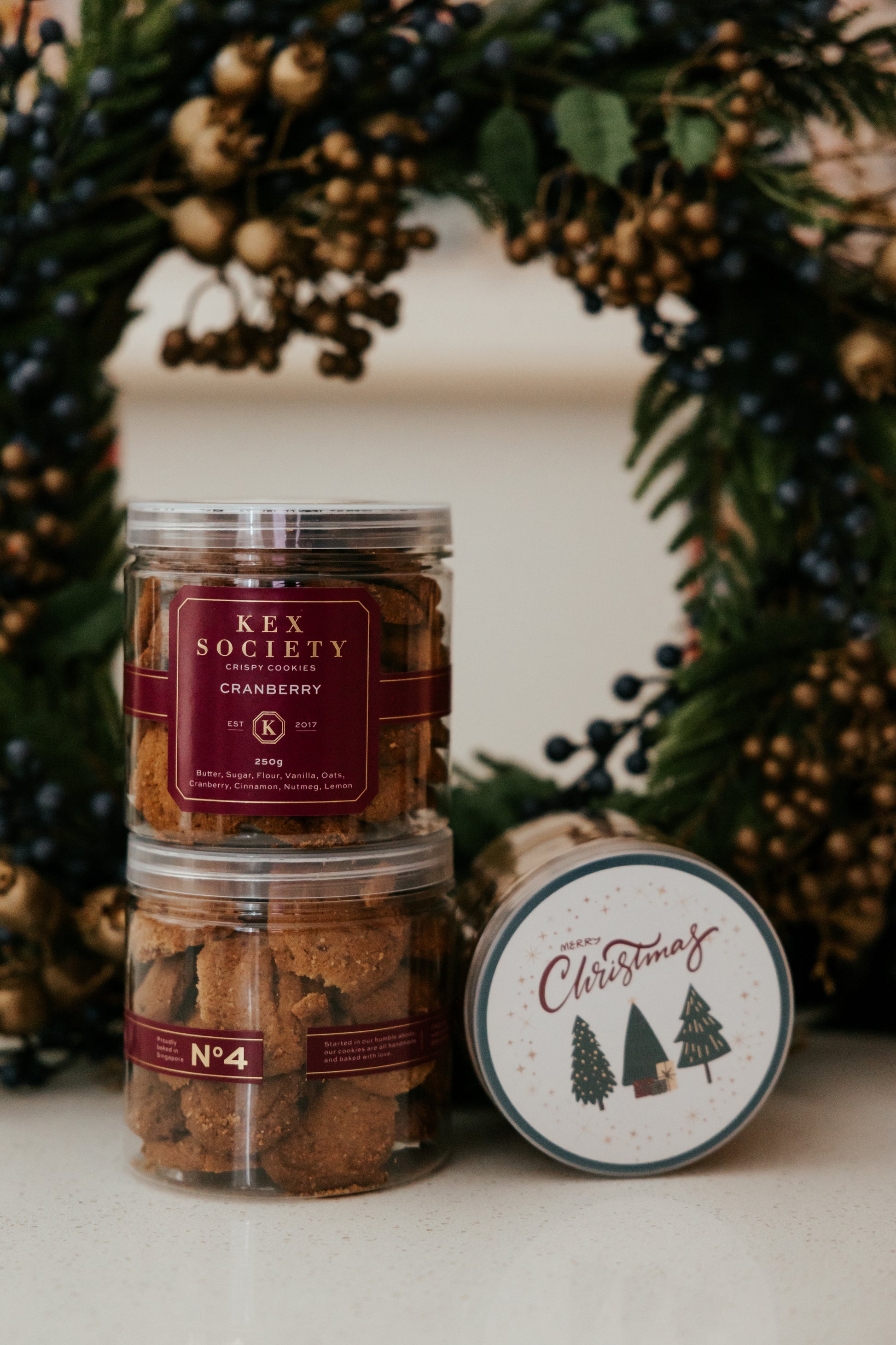 Two jars of KEX Society cranberry cookies with a Christmas-themed label in front of a festive wreath.