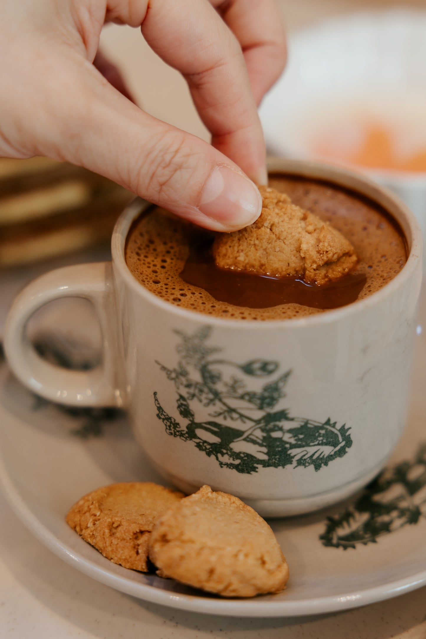 Hand dipping a kex society  better batter cookie into a cup of coffee with a decorative handle and green design.