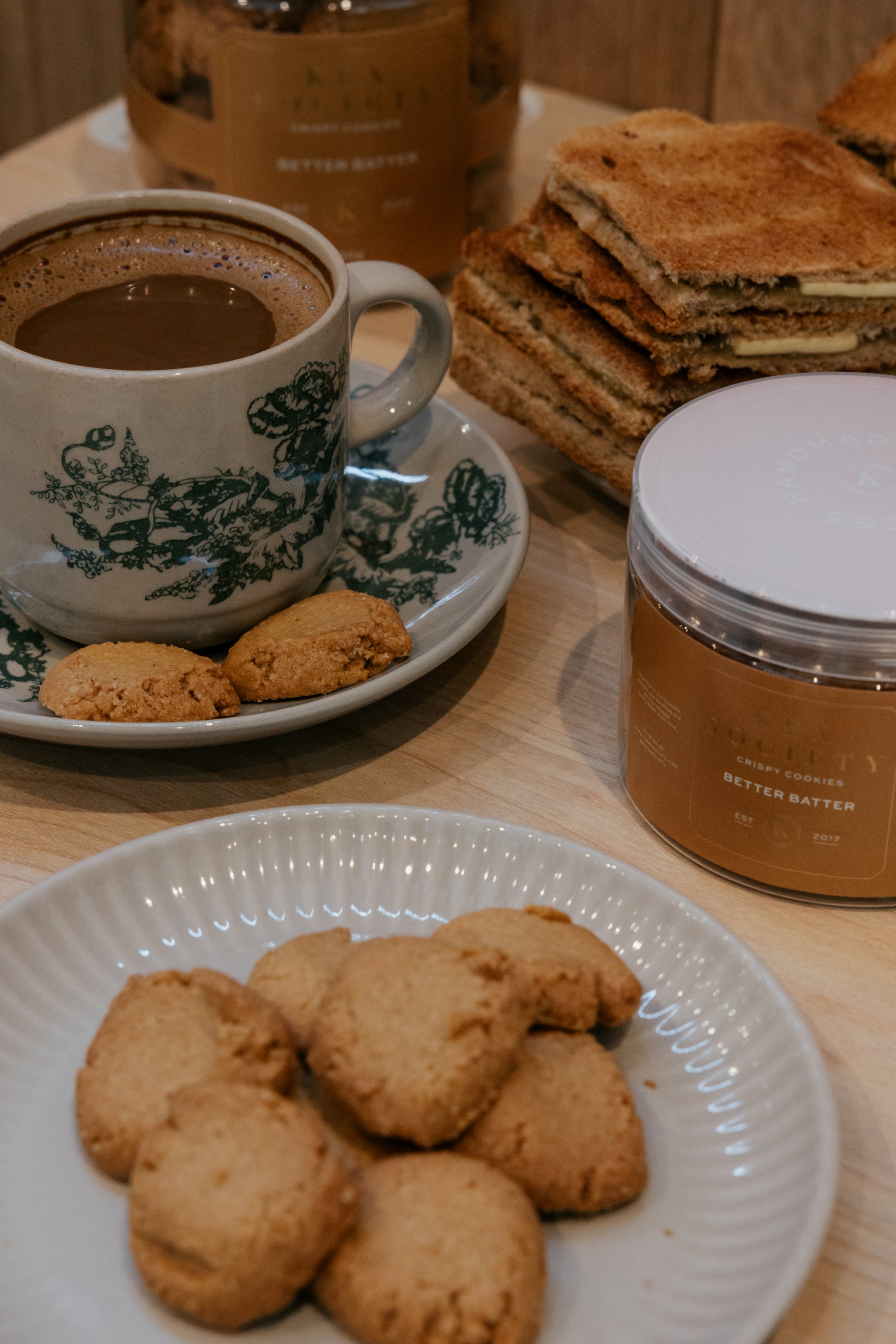 Cup of coffee with kex society better batter cookies on a plate, surrounded by Kex Society cookie bottles and toast.