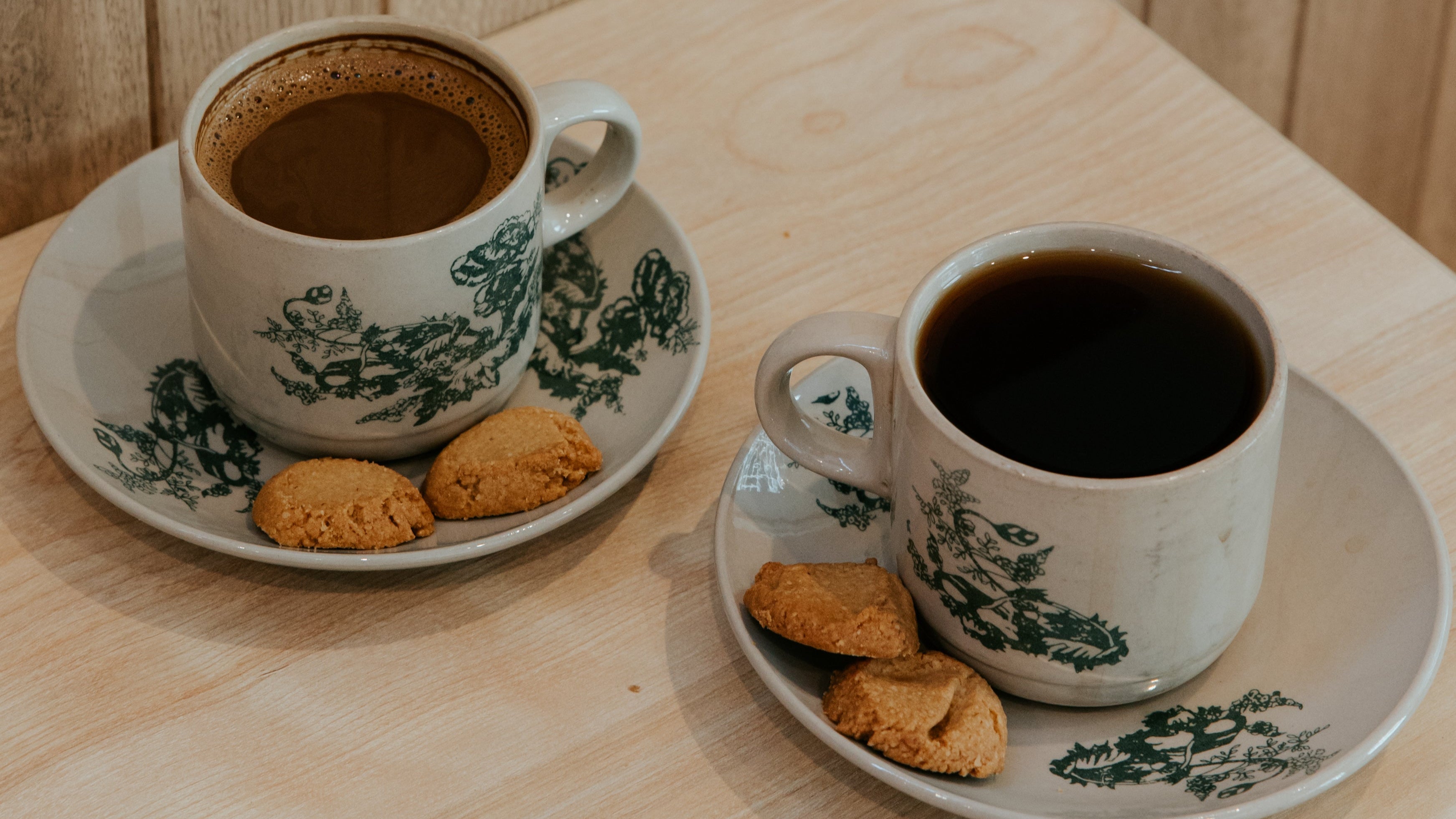 Two cups of coffee with cookies on a wooden table against a wooden paneled wall.
